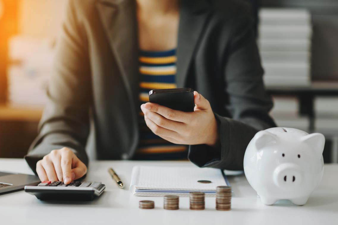 Person sitting at their work desk looking at their phone while using a calculator with a piggy bank and coins on their desk. 