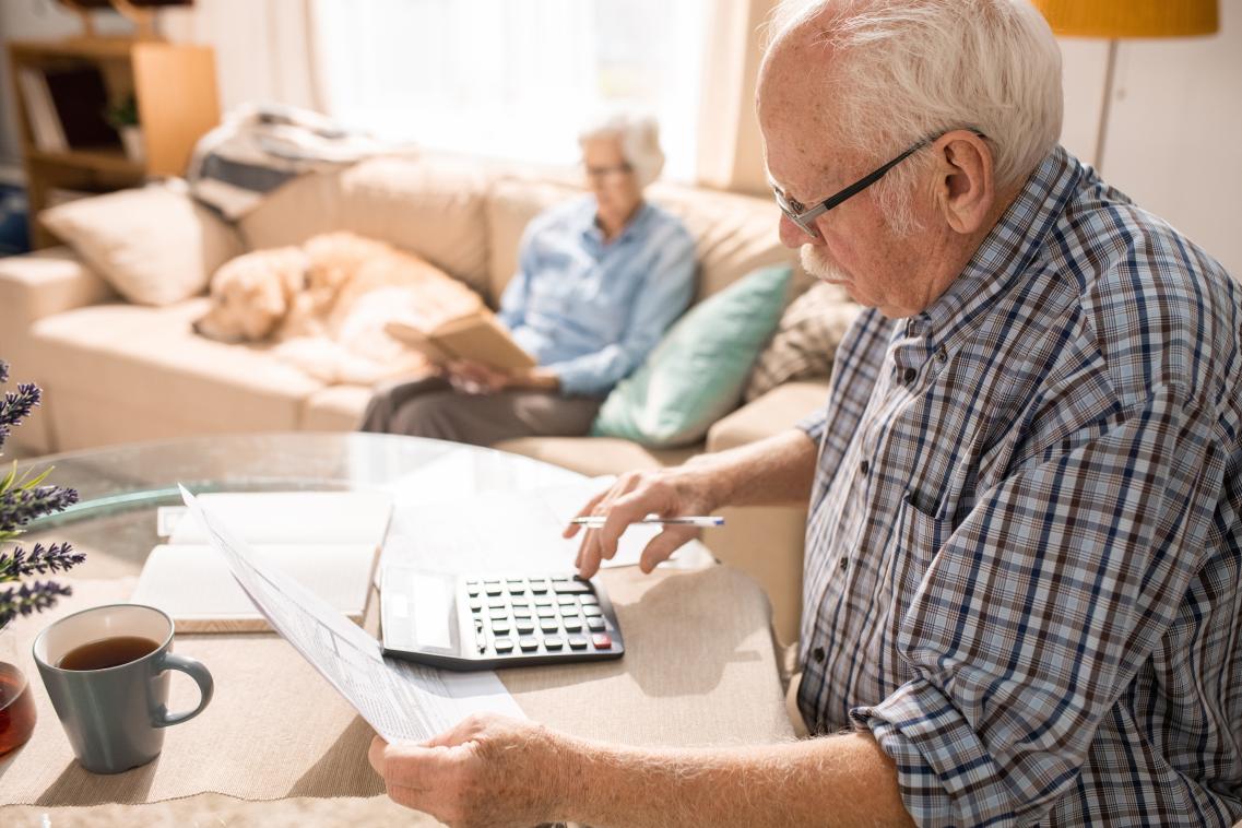 Elderly man using a calculator while sitting at table with elderly woman and pet dog sitting on couch in the background