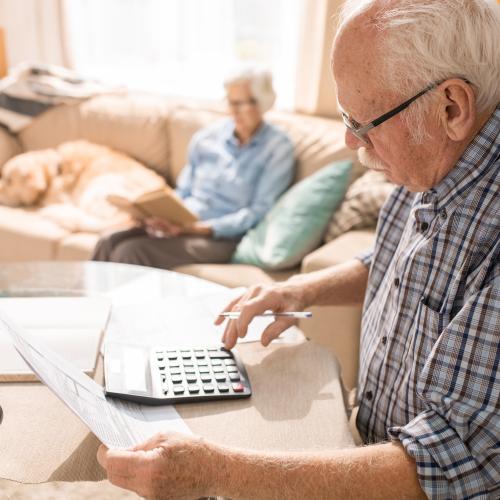 Elderly man using a calculator while sitting at table with elderly woman and pet dog sitting on couch in the background