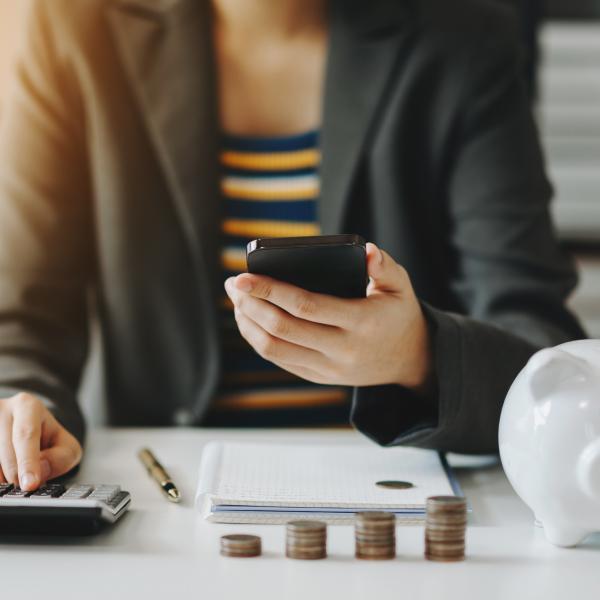 Person sitting at their work desk looking at their phone while using a calculator with a piggy bank and coins on their desk. 